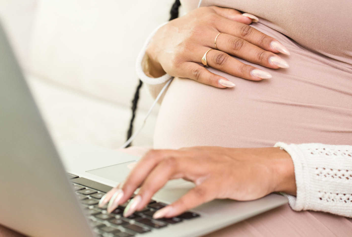 pregnant black woman sitting on bed with upper half proppe on a pillow with one hand on extended belly and the other hand over laptop that is balancing on lap